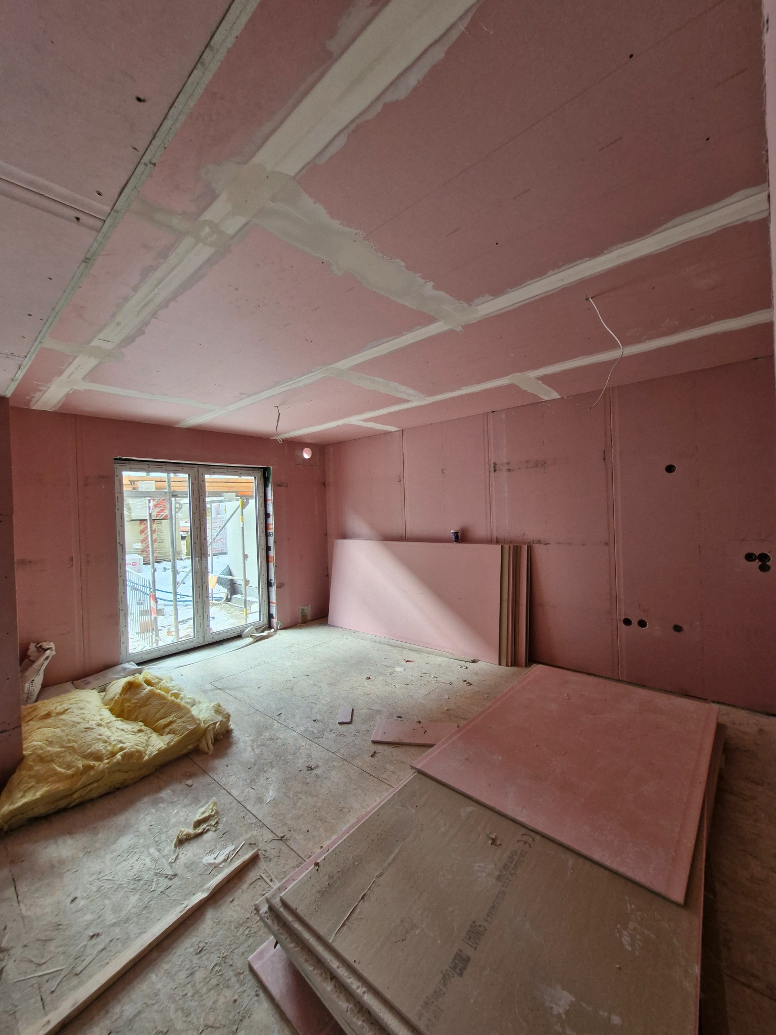 Wnętrze pokoju w trakcie montażu okładziny z płyt w kolorze różowym, widoczne profile, izolacja z wełny mineralnej, oraz stos płyt czekających na montaż.