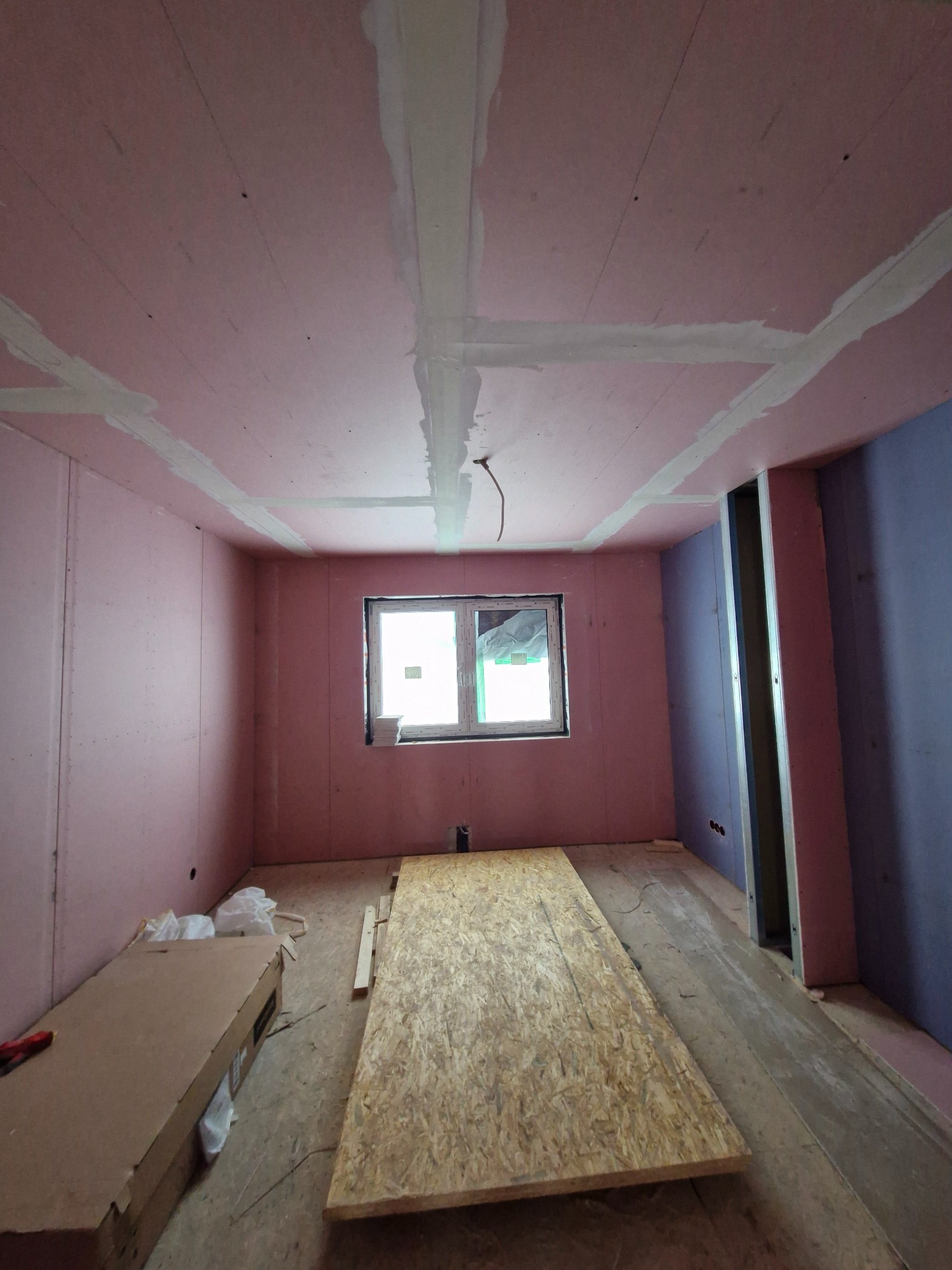 Pokój w trakcie wykańczania: ściany i sufit pokryte różowymi płytami, łączenia szpachlowane, widoczne płyty OSB na podłodze, okno z widokiem na zewnątrz.