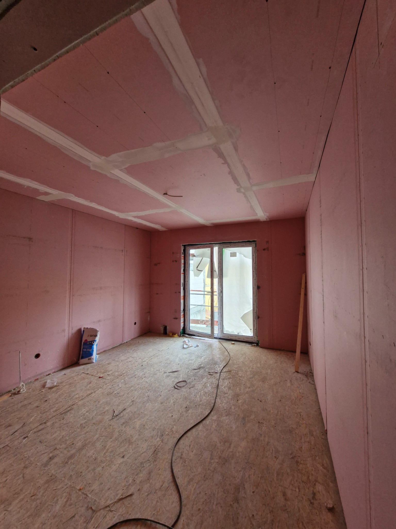 Pokój w trakcie wykańczania: różowe ściany z płyt, szpachlowane łączenia na suficie, surowa podłoga z płyty OSB, okno balkonowe i worek zaprawy na podłodze