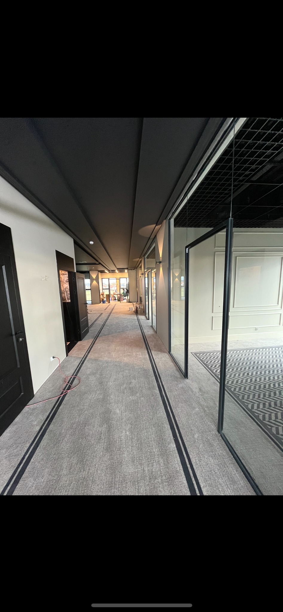 Długi korytarz biurowy w trakcie wykończenia, z szarą wykładziną podłogową z czarnymi paskami, czarnym sufitem kasetonowym i szklanymi ściankami działowymi oddzielającymi pomieszczenia, widoczne...