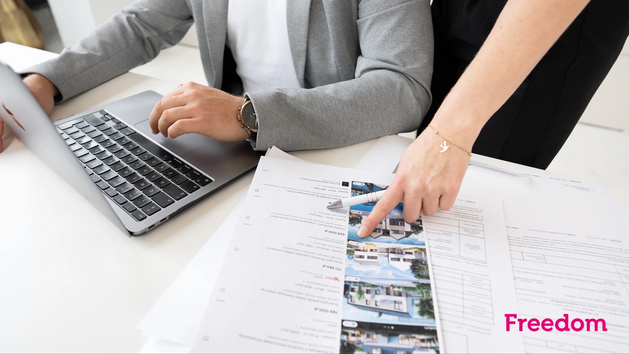 Osoba wskazująca długopisem na zdjęcie domu w ofercie, obok laptop i dokumenty, na nadgarstku bransoletka z motywem ptaka.