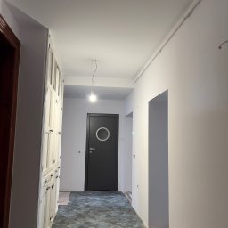 JAR-BUD - Korytarz w trakcie odświeżania, ściany pomalowane na biało, widoczne ślady po instalacjach elektrycznych, podłoga wyłożona płytkami imitującymi kamień, w tle czarne drzwi z okrągłym okienkiem...