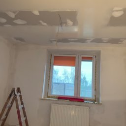 JAR-BUD - Pokój w Raciborzu w trakcie remontu: sufit i ściany po szpachlowaniu, widoczne niedoskonałości i wystające przewody elektryczne, drabina oparta o ścianę, okno z roletą zabezpieczone taśmą malarską.