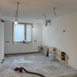 JAR-BUD - Wnętrze w trakcie remontu: surowy beton na podłodze, częściowo wyszpachlowane ściany z wystającymi kablami i rurami, okno zabezpieczone folią, szlifierka do gładzi na podłodze.