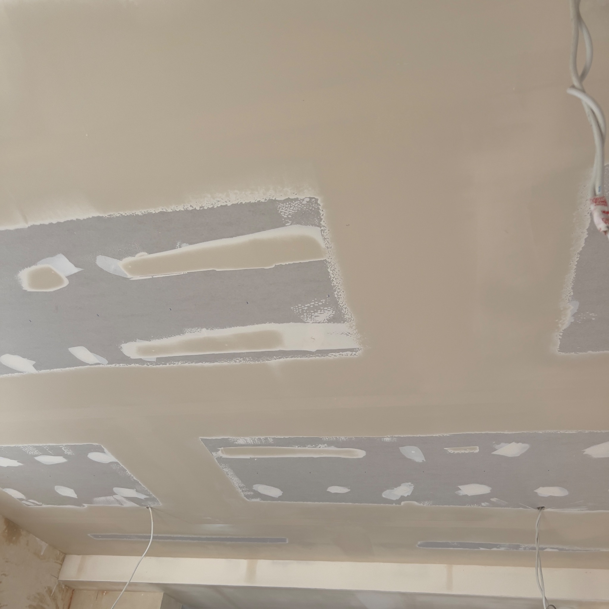 Sufit w trakcie prac wykończeniowych z widocznymi fragmentami płyty kartonowo-gipsowej, świeżo położoną gładzią i wystającymi przewodami elektrycznymi.