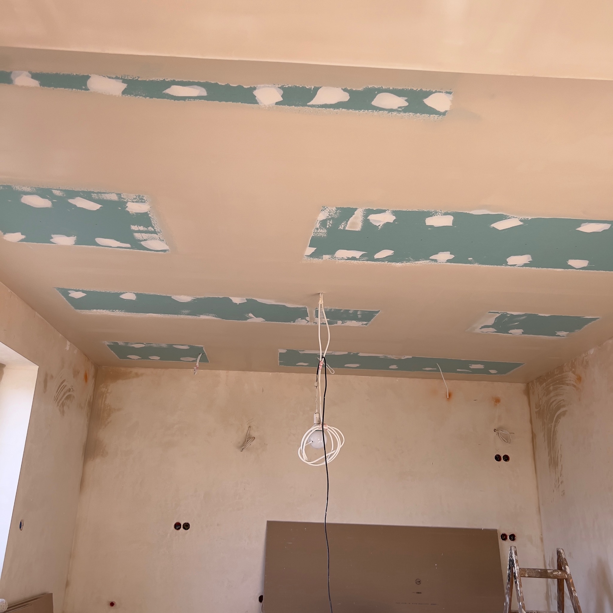 Niedokończony sufit z płyt kartonowo-gipsowych z widocznymi łączeniami i miejscami po szpachlowaniu, surowe ściany i drabina w rogu pokoju.