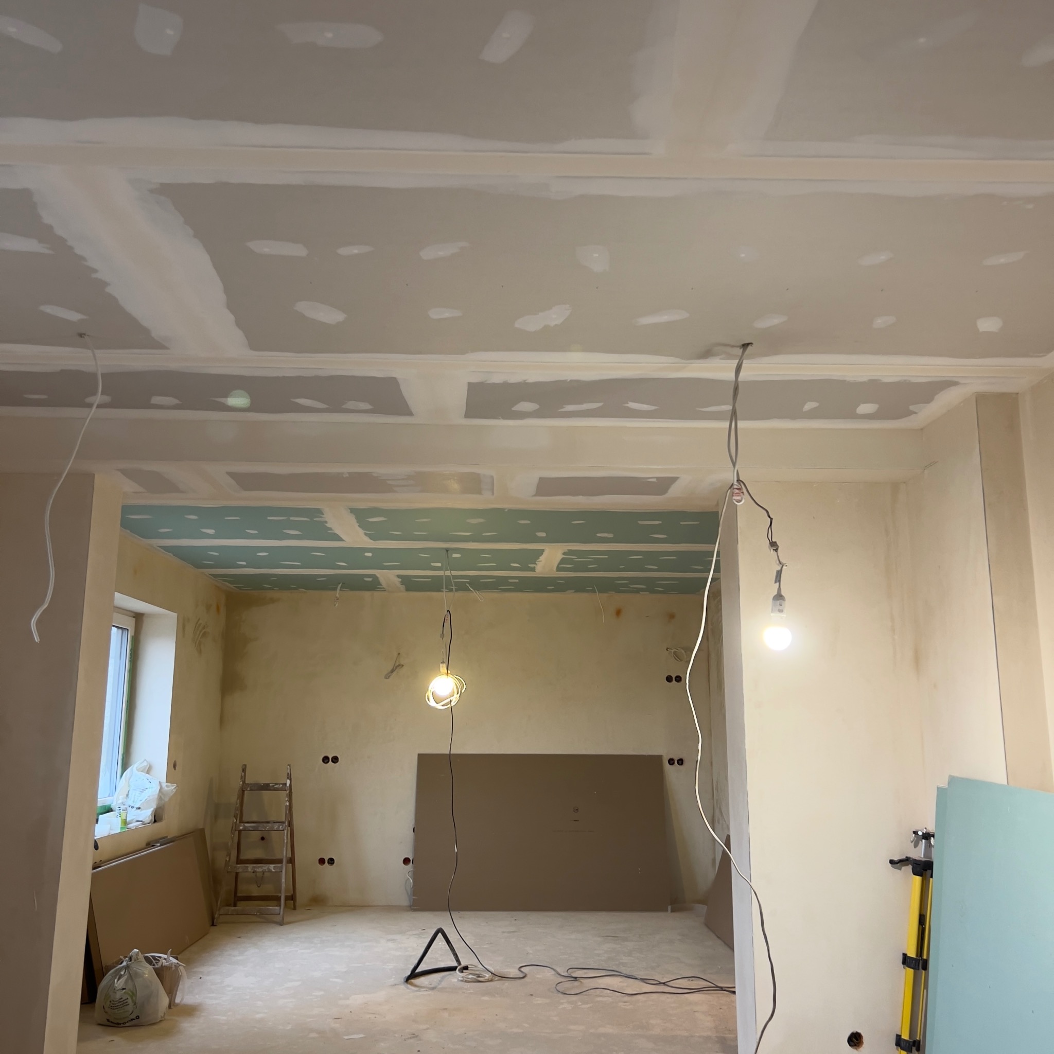 Wnętrze w trakcie prac wykończeniowych: zamontowane płyty kartonowo-gipsowe na suficie i ścianach, widoczne łączenia szpachlowane, prowizoryczne oświetlenie budowlane, drabina i materiały budowlane...