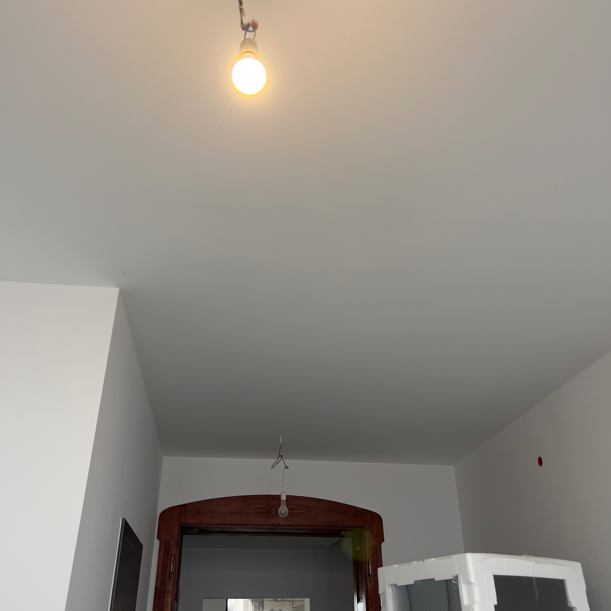 Wnętrze w trakcie remontu, świeżo pomalowane ściany na biało, widoczna żarówka na kablu zwisająca z sufitu i futryna drzwi. Na ścianie widoczna czerwona zaślepka gniazdka.