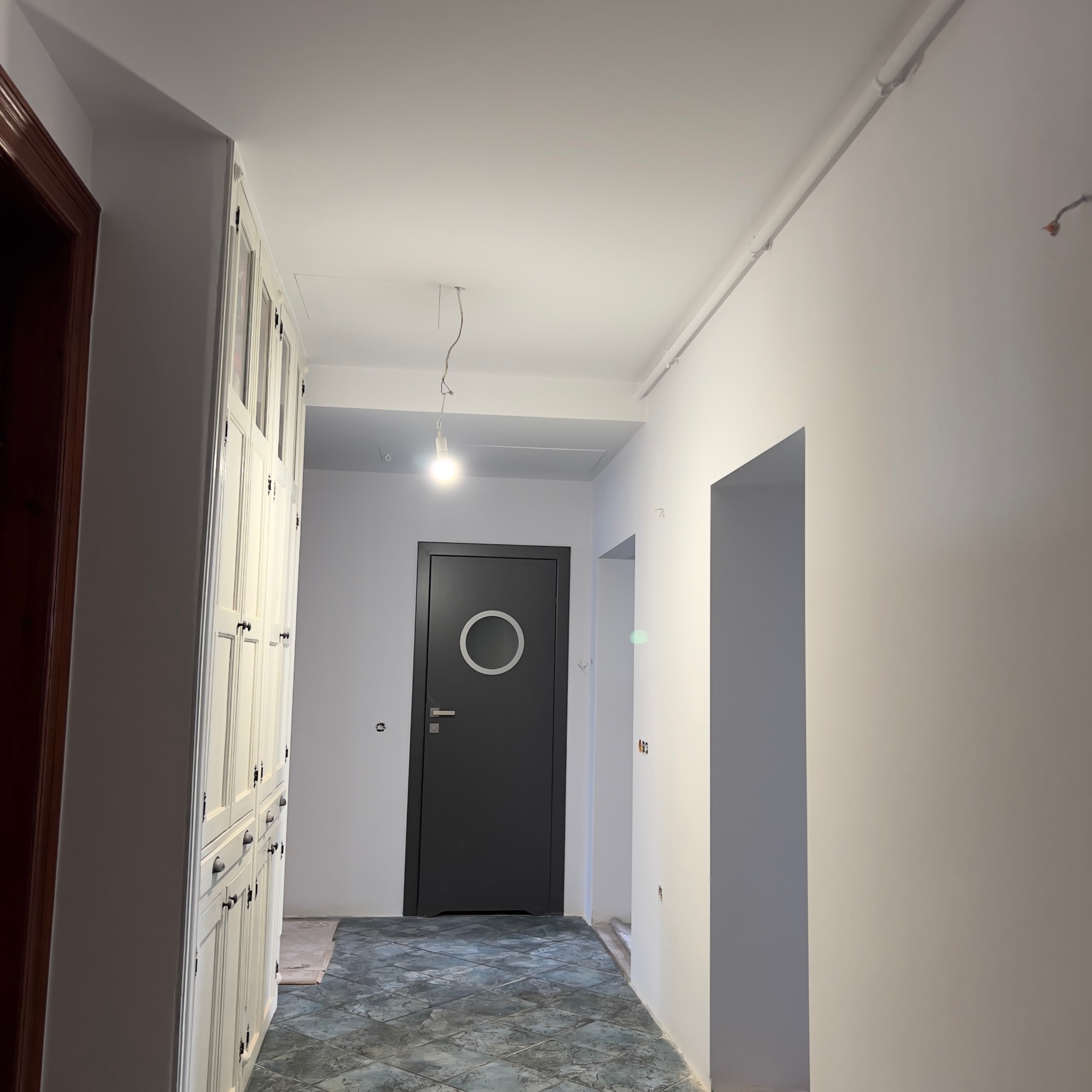Korytarz w trakcie odświeżania, ściany pomalowane na biało, widoczne ślady po instalacjach elektrycznych, podłoga wyłożona płytkami imitującymi kamień, w tle czarne drzwi z okrągłym okienkiem...