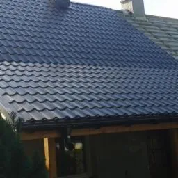 Ciemny dach z blachodachówki modułowej z zamontowanym systemem rynnowym, widok z dołu, fragment elewacji budynku w tle.