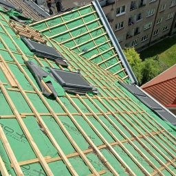 Widok z góry na konstrukcję dachu pokrytą zieloną membraną, z zamontowanymi oknami dachowymi i drewnianą łatą przygotowaną pod pokrycie dachowe. W tle widoczny budynek mieszkalny.