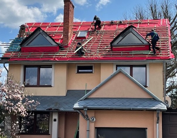 Prace dekarskie na dachu dwupiętrowego domu z dwoma lukarnami trójkątnymi, widoczne rusztowania i ekipa montująca pokrycie na czerwonej membranie.