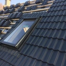 Częściowy widok dachu z zamontowanym oknem dachowym i ułożonymi dachówkami karpiówkami w kolorze grafitowym, widoczne elementy więźby dachowej w trakcie prac dekarskich.