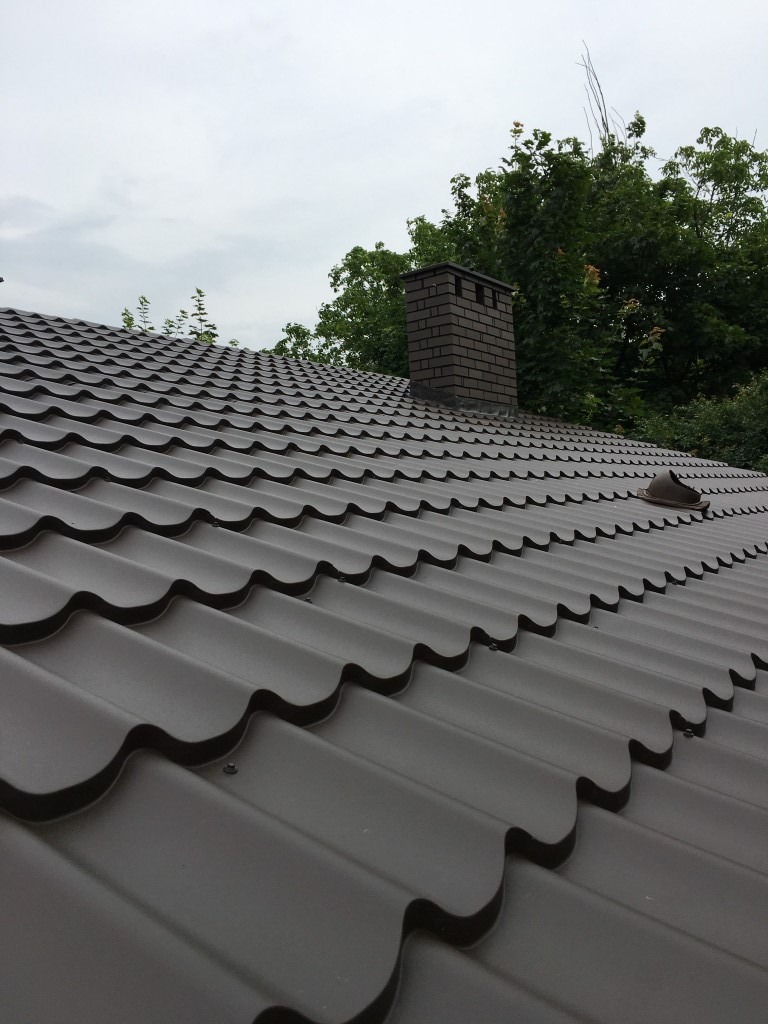 Ciemnobrązowe pokrycie dachowe z blachy trapezowej w kształcie fali, widoczne z niskiego kąta, z kominem z cegły w tle i zielenią drzew.