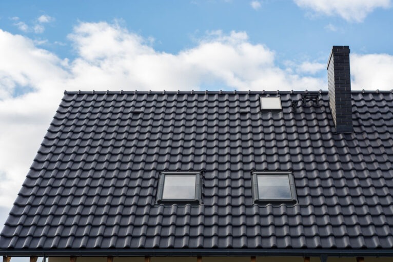 Nowy dach z ciemnoszarej dachówki ceramicznej z trzema oknami dachowymi i kominem z cegły na tle błękitnego nieba z chmurami.