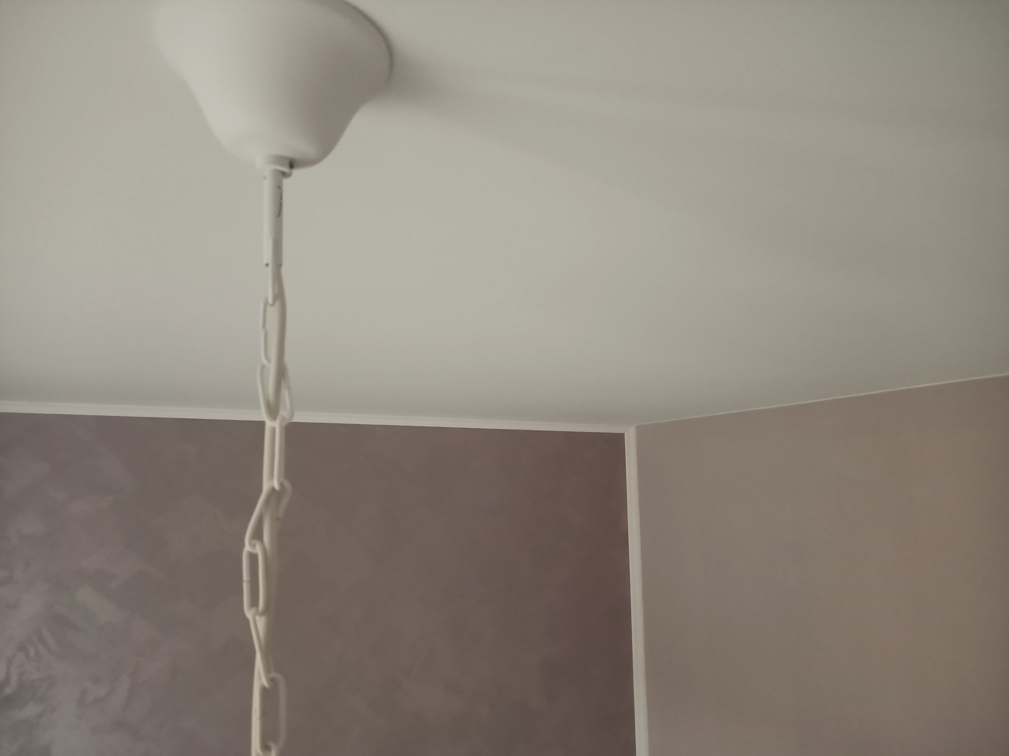 Biała lampa sufitowa z łańcuchem na tle ściany pomalowanej na dwa odcienie szarości, widoczny fragment sufitu.