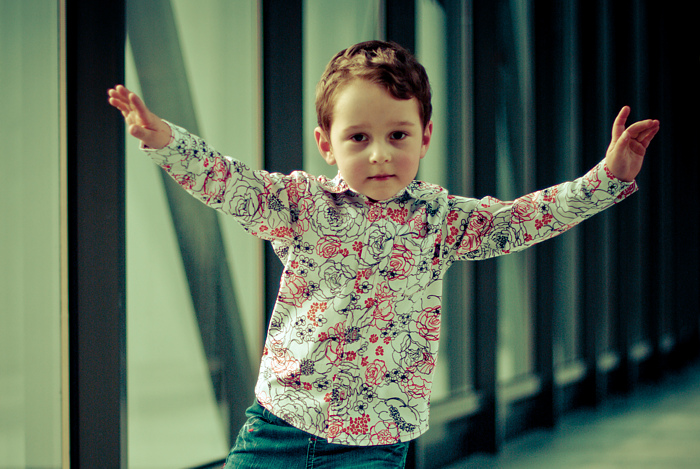 Chłopiec w koszuli w kwiaty, z rozłożonymi rękami, prezentuje kolekcję odzieży dziecięcej producenta z Karsibora na tle nowoczesnego wnętrza z pionowymi listwami.