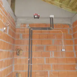 Instalacja elektryczna w surowym wnętrzu: przewody, puszki, rury w ścianach z cegły dziurawki, widoczne elementy drewnianej konstrukcji dachu i prowizoryczne oświetlenie.