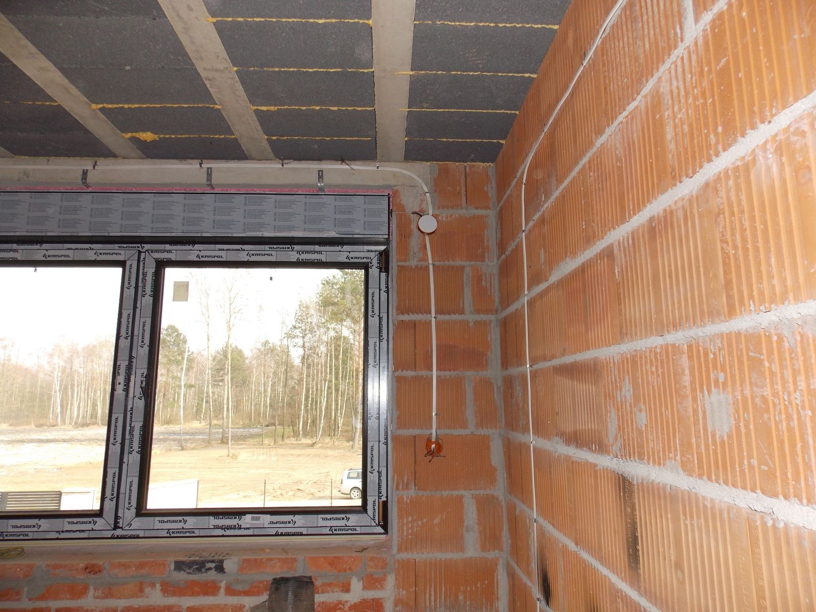 Surowy mur z instalacją elektryczną: przewody prowadzone po ścianie, puszki pod oświetlenie. Widok przez okno na krajobraz. Budynek w trakcie budowy.