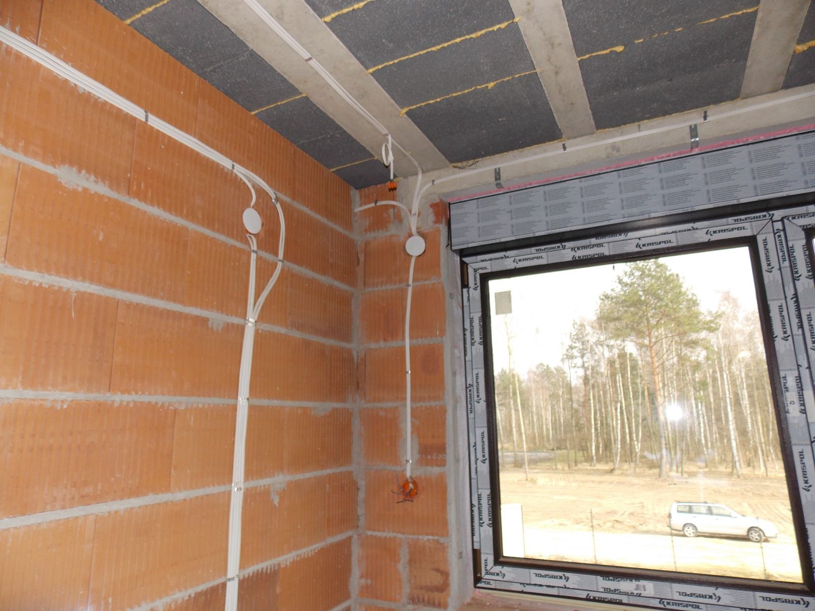 Surowy mur z instalacją elektryczną: kable w białych listwach, puszki pod gniazdka. W tle okno z widokiem na las i auto. Instalacja w trakcie realizacji.