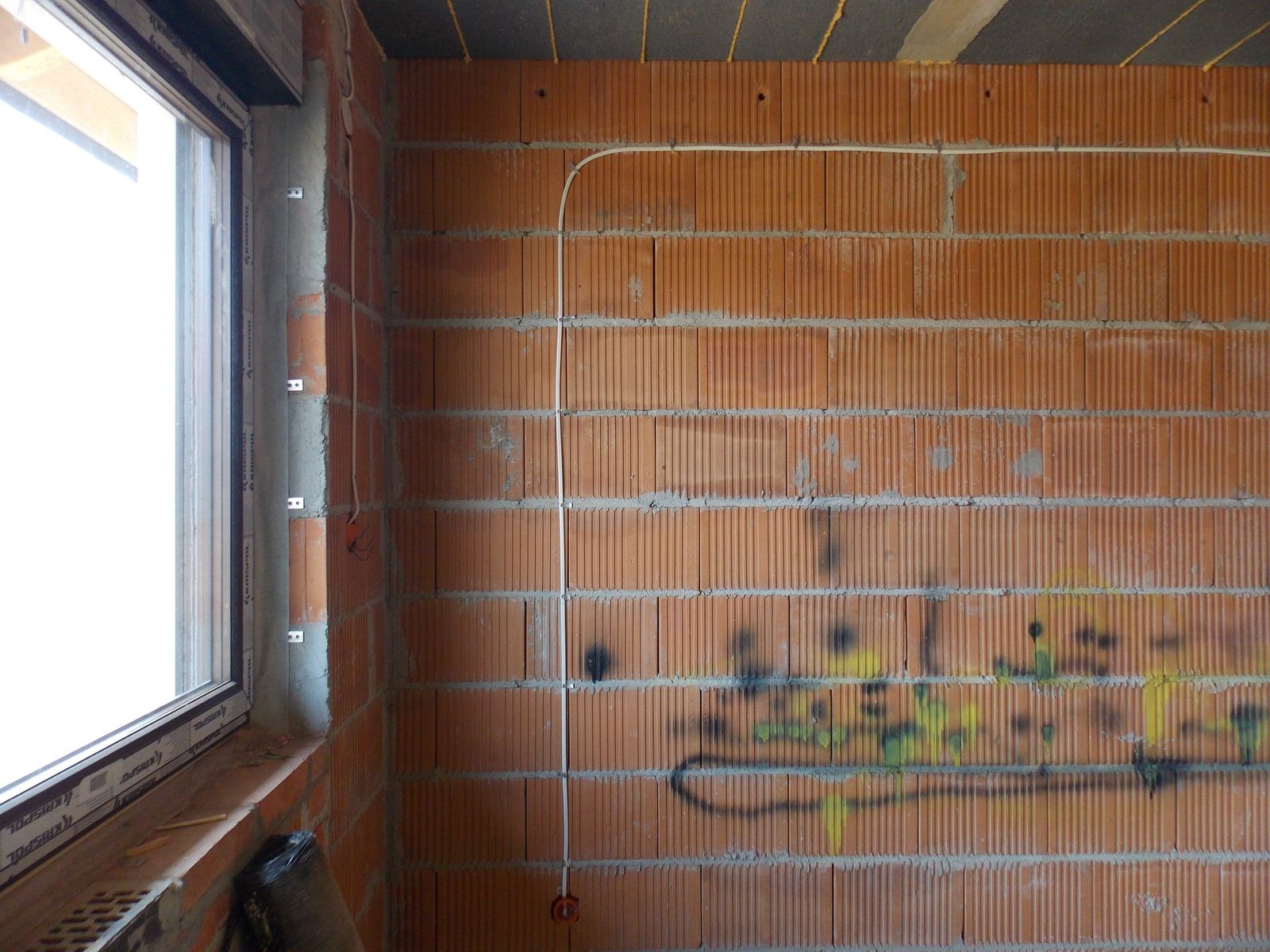 Biały przewód elektryczny poprowadzony po czerwonej ścianie z cegły dziurawki w surowym stanie, widoczne uchwyty mocujące i ślady po graffiti.