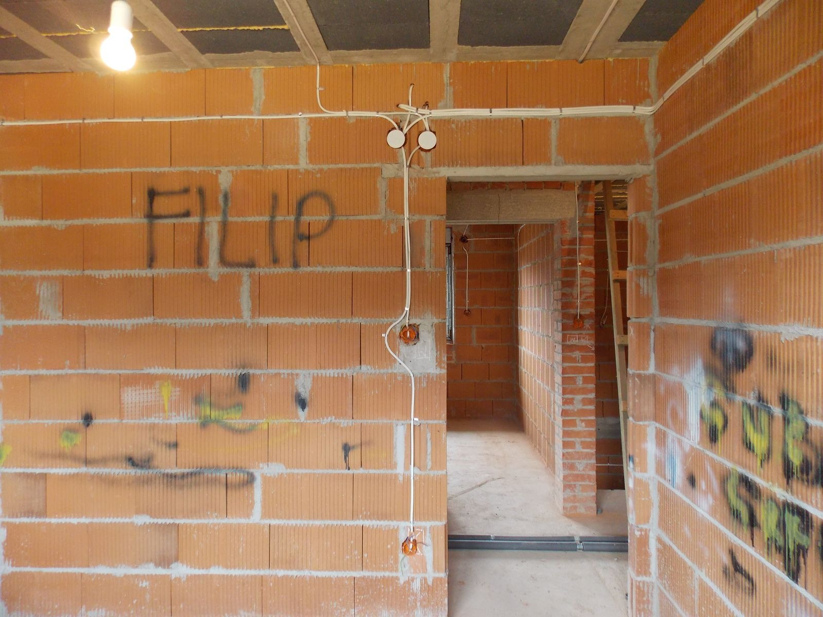 Wewnętrzna instalacja elektryczna w surowym budynku z cegły. Widoczne przewody, puszki elektryczne i żarówka. Ściana z napisem 'FILIP' i śladami farby.