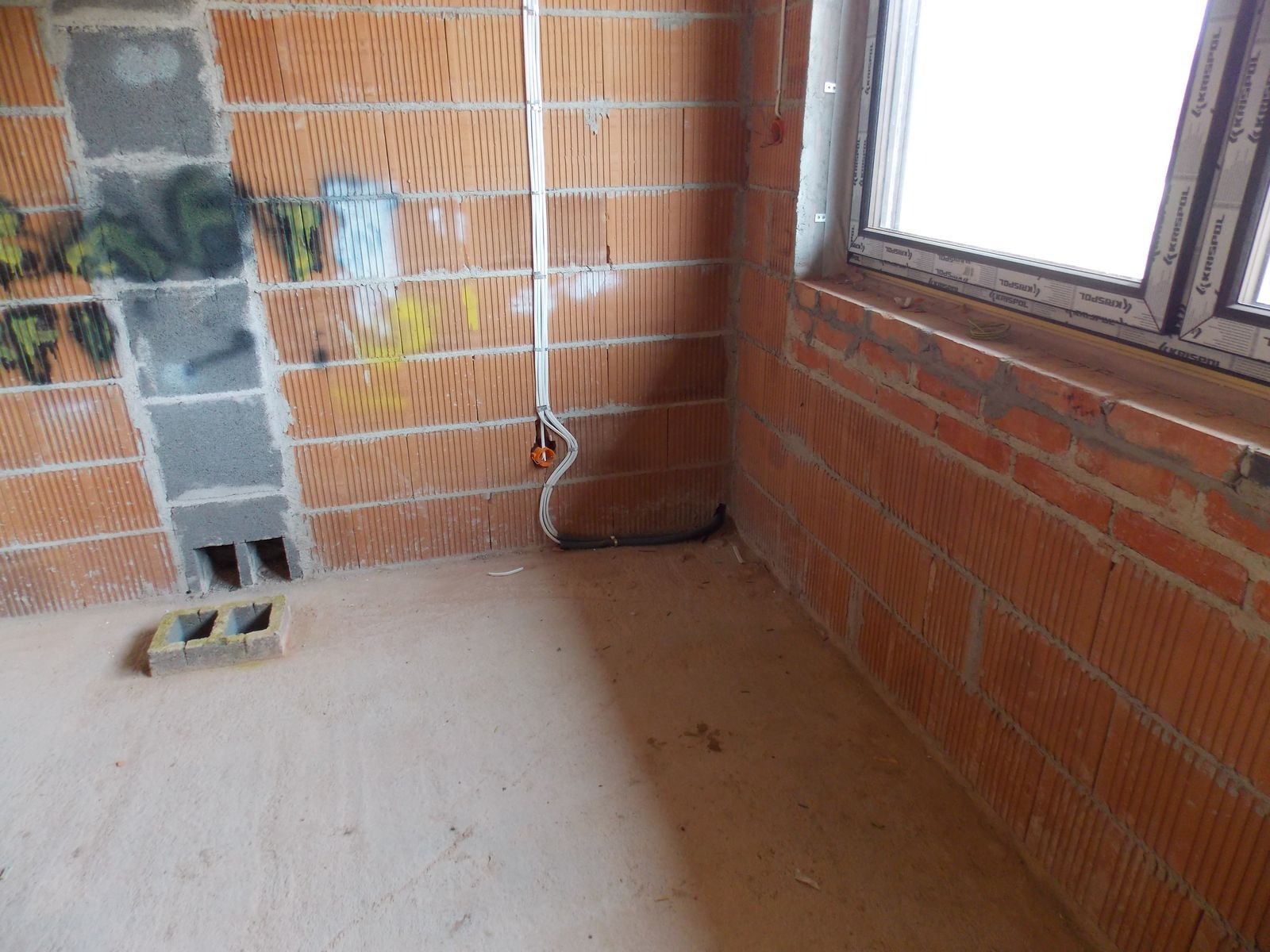 Surowy mur z instalacją elektryczną wewnątrz budynku, widoczne przewody i puszka. W tle okno. Ściany z cegły dziurawki, podłoga betonowa.