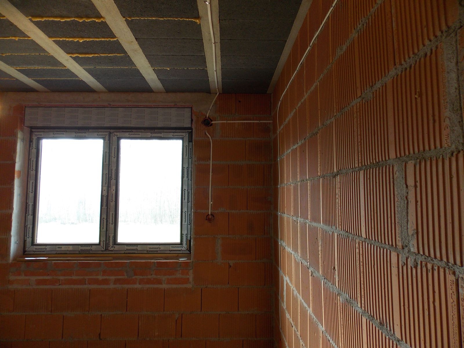Wnętrze w trakcie budowy: ściany z czerwonej cegły, okno z roletą i widoczne przewody elektryczne w peszlu, prowadzone po ścianie do puszek instalacyjnych.