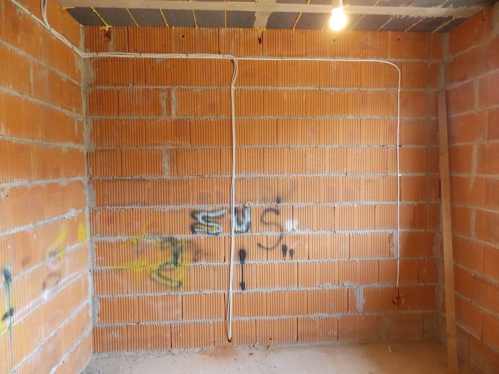 Wnętrze w trakcie budowy z widoczną instalacją elektryczną poprowadzoną po ceglanej ścianie z napisem. Żarówka oświetla surowe pomieszczenie.