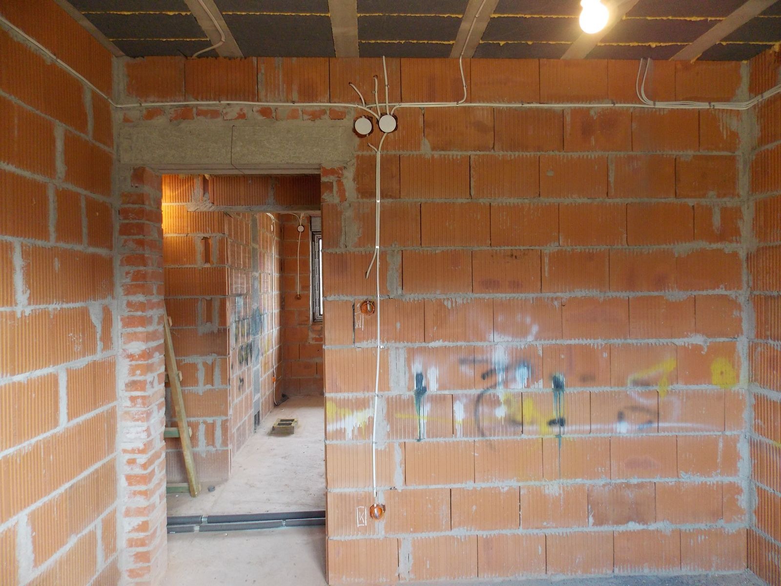 Surowy mur z cegły dziurawki z widoczną instalacją elektryczną: kable w peszlach i puszki pod gniazdka. W tle widoczne kolejne pomieszczenie w stanie deweloperskim.