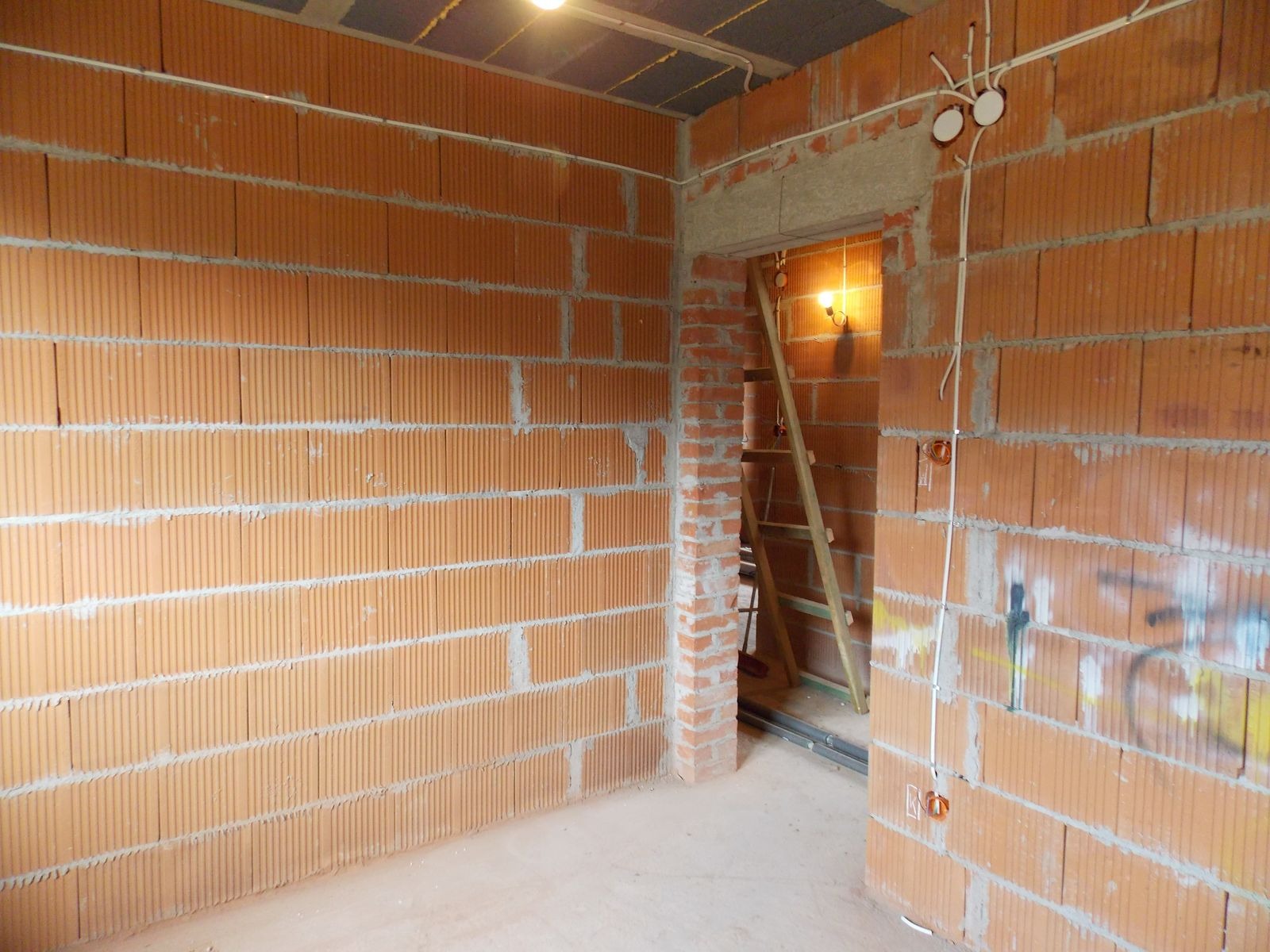 Surowy mur z instalacją elektryczną w trakcie montażu. Widoczne przewody, puszki i fragment drabiny w przejściu. Oświetlenie sufitowe i brak tynków.