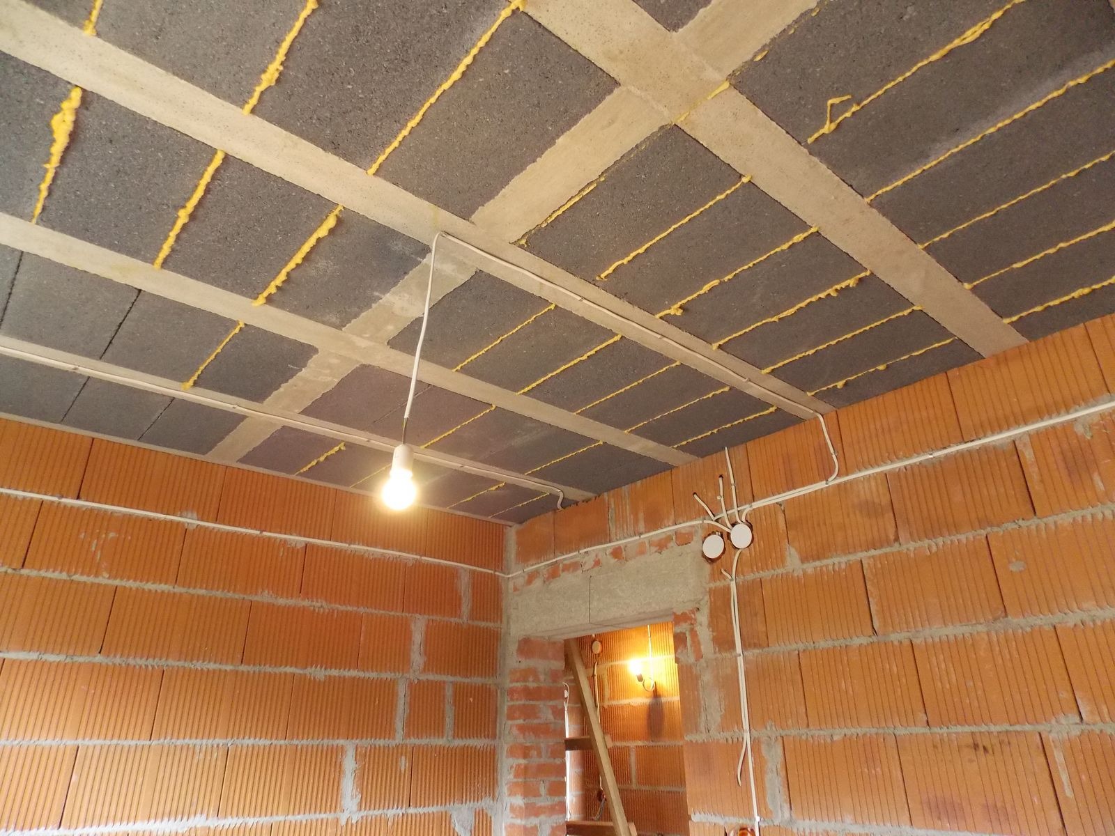 Surowy stan: instalacja elektryczna w budynku z cegły, widoczne kable prowadzone po suficie z pustaków i ścianach, punkt oświetleniowy z żarówką.