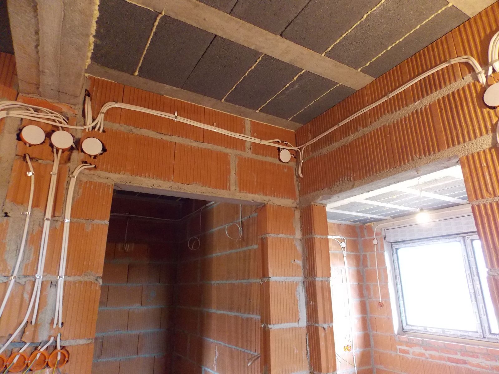Surowy stan: instalacja elektryczna w budynku z cegły kratówki. Widoczne kable, puszki instalacyjne, strop z izolacją. Oświetlenie tymczasowe.