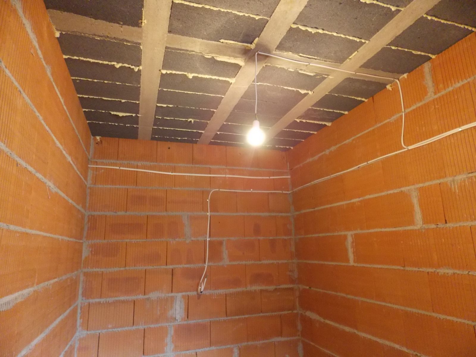 Wnętrze w trakcie budowy: surowa cegła, strop z izolacją i prowizoryczne oświetlenie żarówką. Widoczne przewody elektryczne poprowadzone po ścianach.
