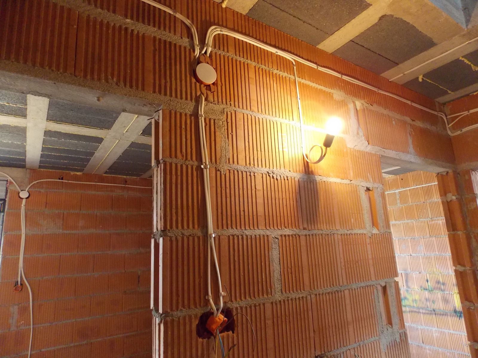 Instalacja elektryczna w surowym budynku z cegły, widoczne kable, puszki i oświetlenie tymczasowe. Prace instalacyjne w trakcie realizacji.