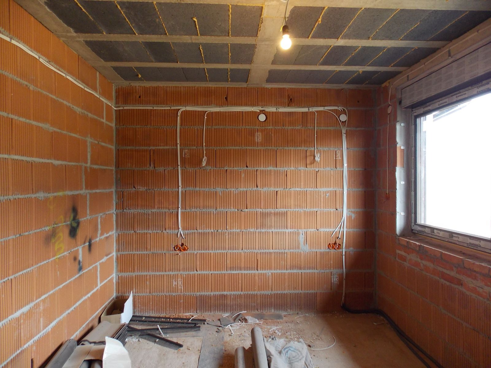 Wnętrze w trakcie prac: widoczne ściany z cegły dziurawki, rozprowadzona instalacja elektryczna w białych rurkach, surowy sufit z widocznymi pustakami stropowymi.