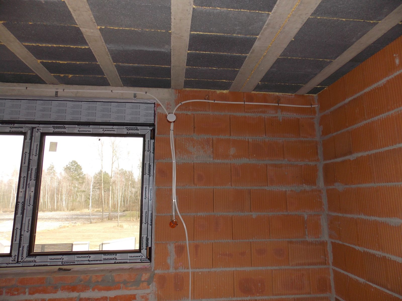 Surowy stan: instalacja elektryczna w peszlu przy oknie, widoczna puszka instalacyjna. Murowana ściana z cegły, strop z pustaków. Stan deweloperski.