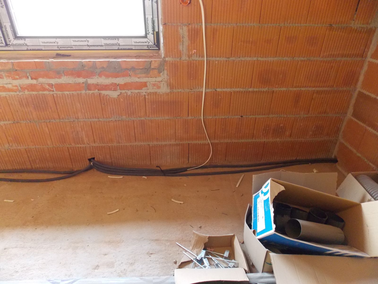 Wewnętrzny widok surowego budynku z instalacją elektryczną przy oknie, kable ułożone wzdłuż ściany z cegły dziurawki i kartony z elementami instalacji.