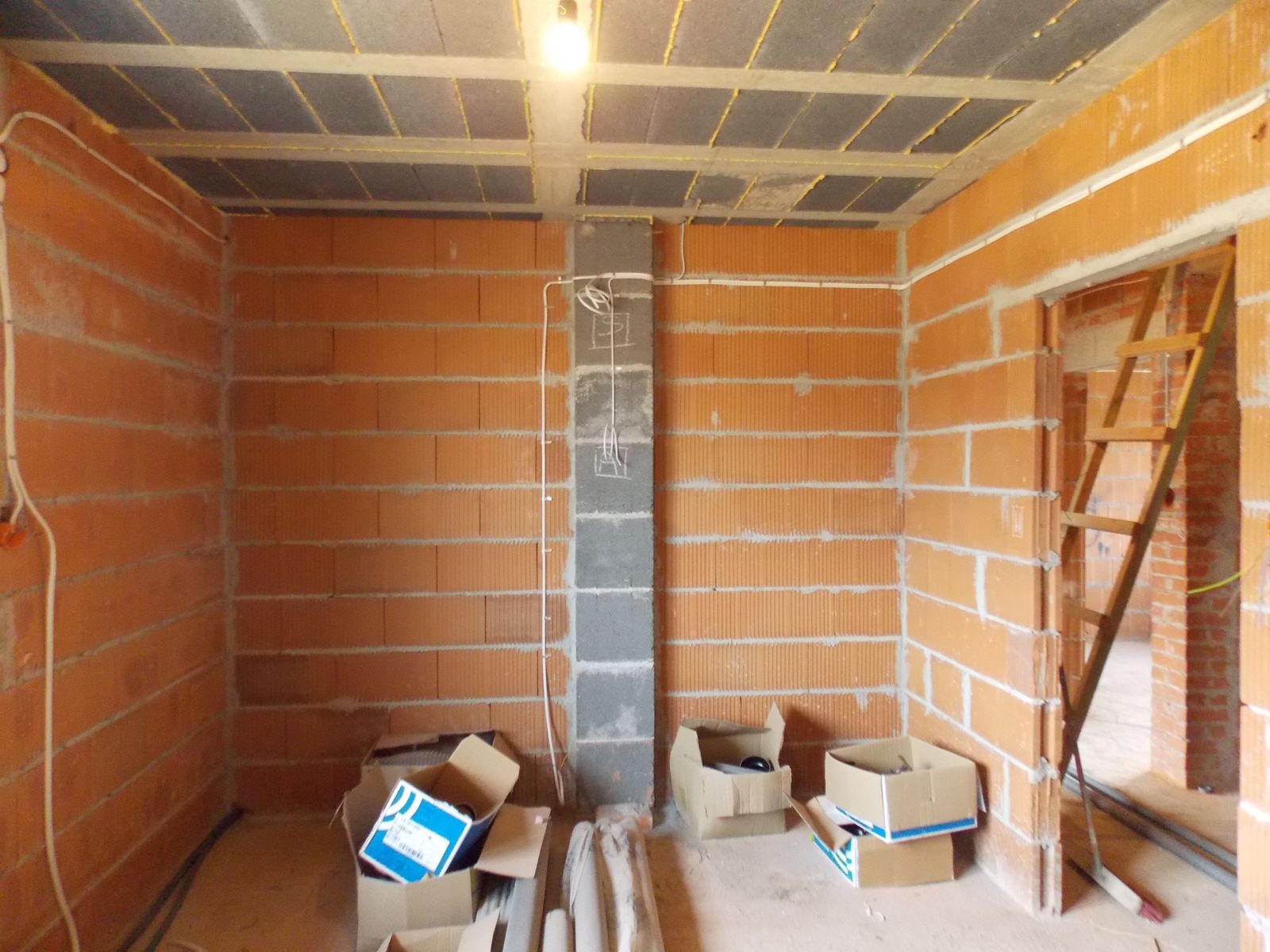 Surowy stan: instalacja elektryczna w ceglanym pokoju z widocznymi kablami, puszkami i betonowym filarem. Kartony na podłodze, drabina w przejściu.