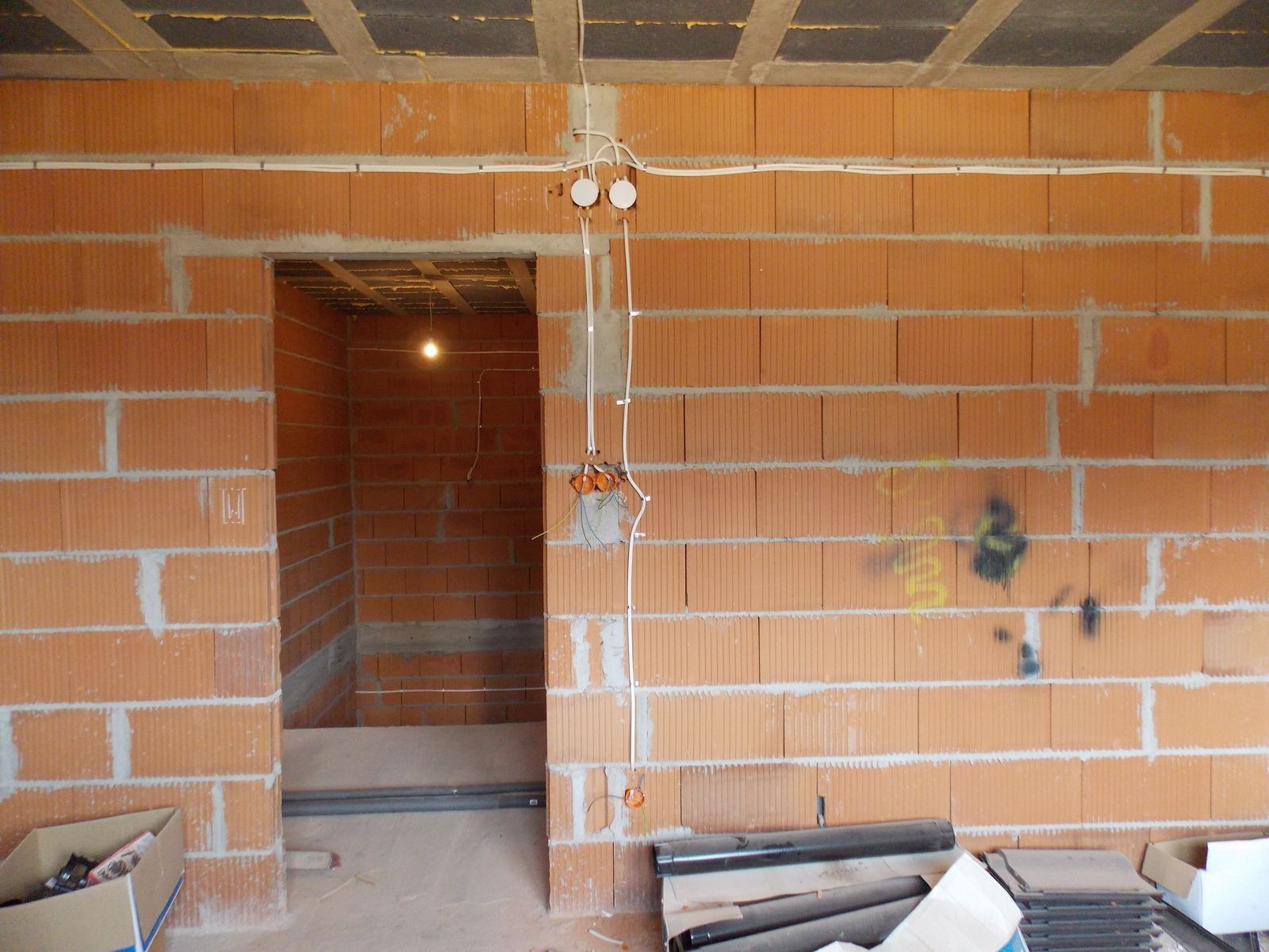 Surowa ściana z cegły z widoczną instalacją elektryczną: puszki, kable i przewody. W tle otwór drzwiowy z widoczną instalacją wewnątrz pomieszczenia.