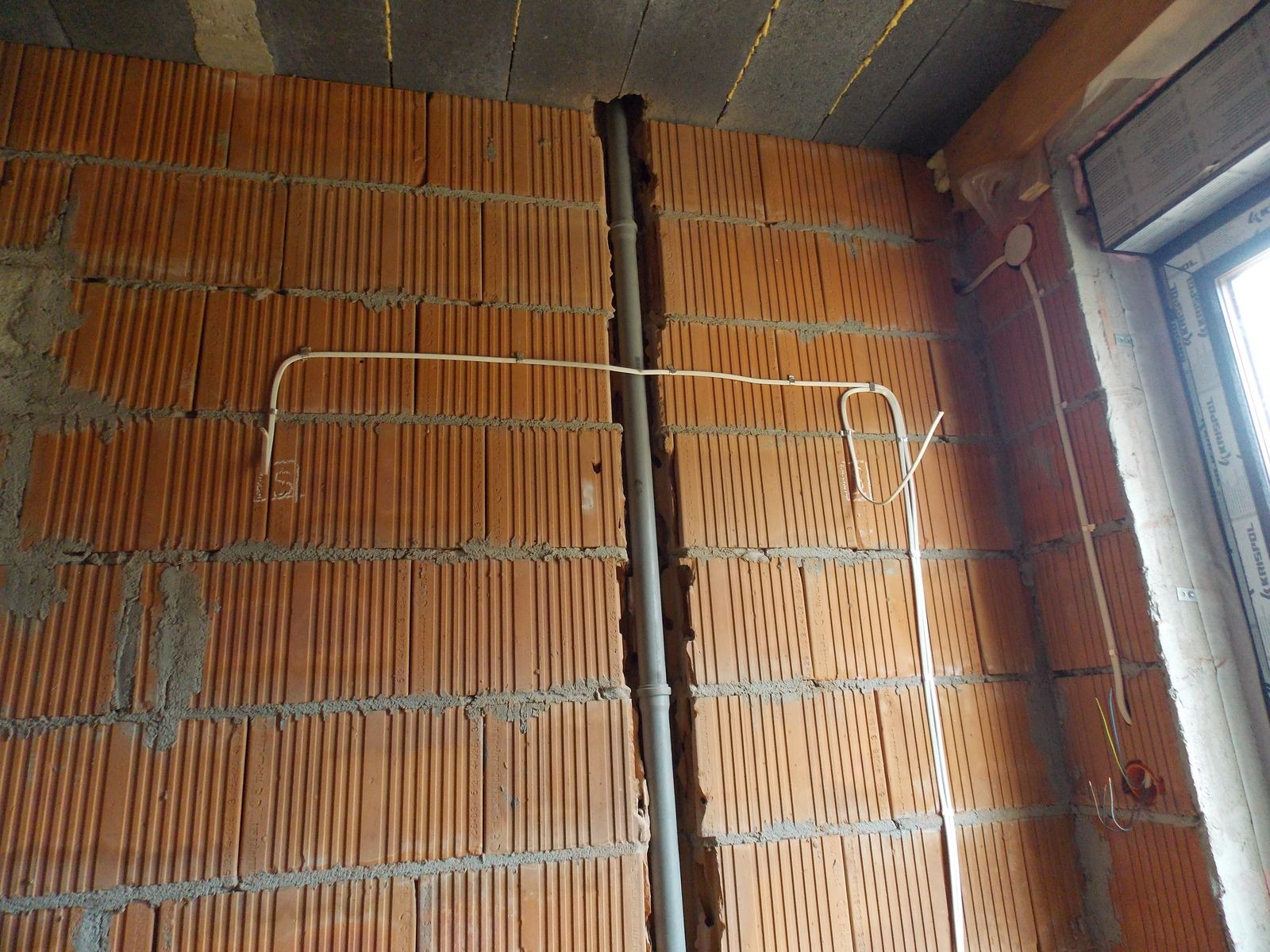 Ceglana ściana z bruzdą, w której umieszczono szarą rurę. Widoczne białe przewody elektryczne, częściowo zamontowane, prowadzone po ścianie w poziomie i pionie.