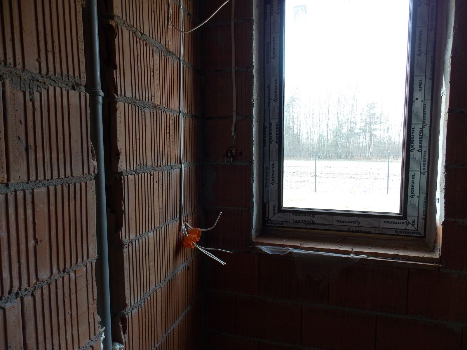 Wnętrze w trakcie budowy, widoczne instalacje elektryczne w ścianie z cegły, puszki elektryczne z przewodami oraz szara rura instalacyjna. Okno z widokiem na zewnątrz.