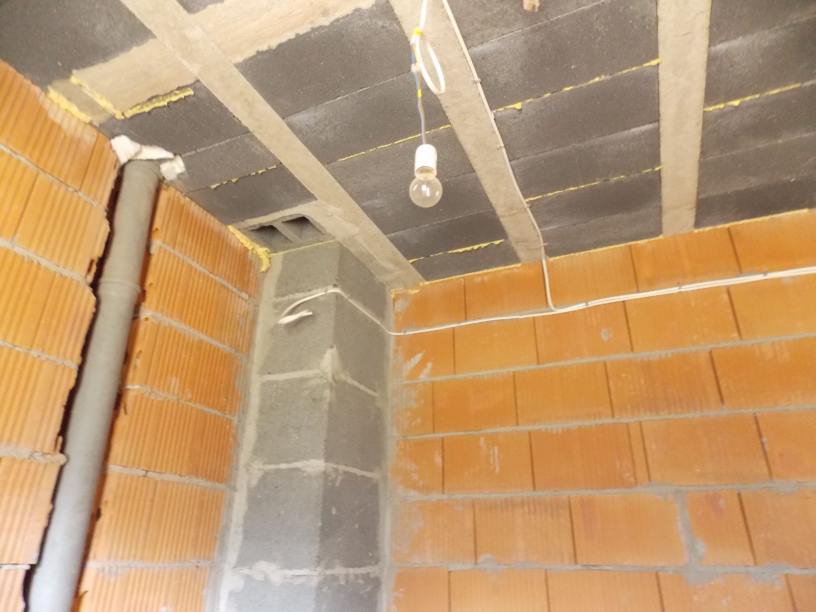 Wnętrze w trakcie budowy: surowy strop z belkami, pomarańczowe cegły, szara rura pionowa, prowizoryczne oświetlenie żarówką na kablu i białe kable elektryczne.