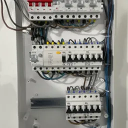 Wnętrze białej skrzynki rozdzielczej z zamontowanymi bezpiecznikami różnych producentów (Eaton, Ideal, ETI) i kolorowymi złączkami kablowymi, widoczne przewody w różnych kolorach i grubościach.