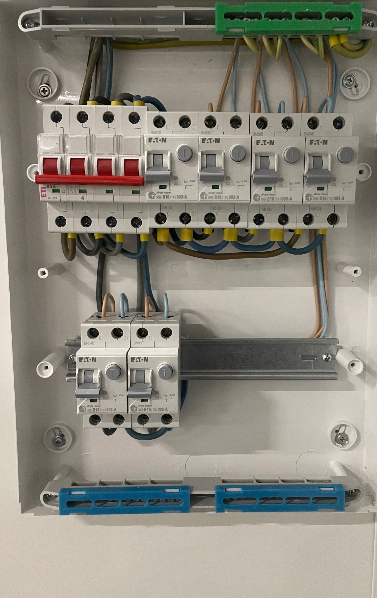 Rozdzielnica elektryczna z zainstalowanymi bezpiecznikami EATON, widoczne przewody z żółtymi i niebieskimi oznaczeniami, szyna montażowa oraz niebieskie elementy mocujące. Jeden z bezpieczników ma...