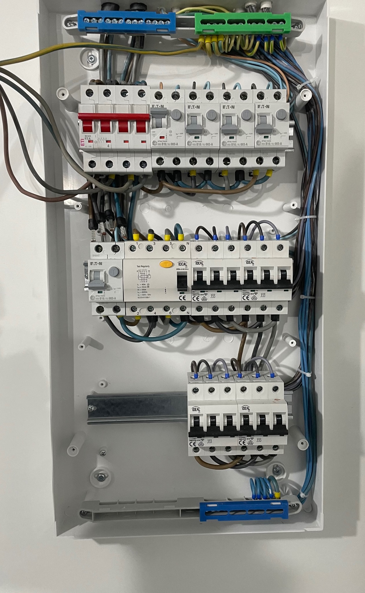 Wnętrze białej skrzynki rozdzielczej z zamontowanymi bezpiecznikami różnych producentów (Eaton, Ideal, ETI) i kolorowymi złączkami kablowymi, widoczne przewody w różnych kolorach i grubościach.