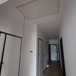 Przemysław Filipiak - Korytarz w trakcie wykończenia, widoczna otwarta klapa rewizyjna w suficie z wystającym elementem podtrzymującym, świeżo pomalowane białe ściany i ciemne obramowania drzwi.