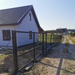Nowo zainstalowane, czarne ogrodzenie panelowe z siatki z betonową podmurówką, widoczne na tle białego domu z ciemną dachówką i polnej drogi.
