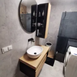 Nowoczesna łazienka z owalną umywalką nablatową na drewnianej szafce, okrągłym lustrem i szarą płytką imitującą beton na ścianach, z widoczną pralką i czarnym grzejnikiem drabinkowym.