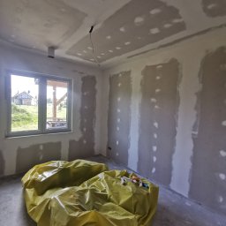 Przemysław Filipiak - Pokój w trakcie prac wykończeniowych: ściany i sufit pokryte wstępną warstwą gładzi, podłoga zabezpieczona żółtą folią malarską, widoczne narzędzia malarskie na folii, okno z widokiem na budynek...